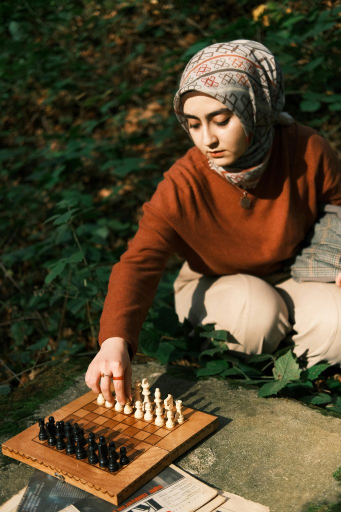 A young woman in a hijab plays chess outdoors, focusing on the game in a natural setting.