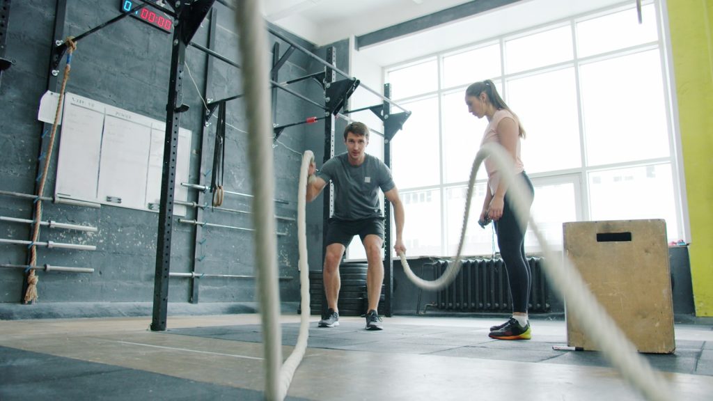Man and woman exercising with battle ropes indoors.